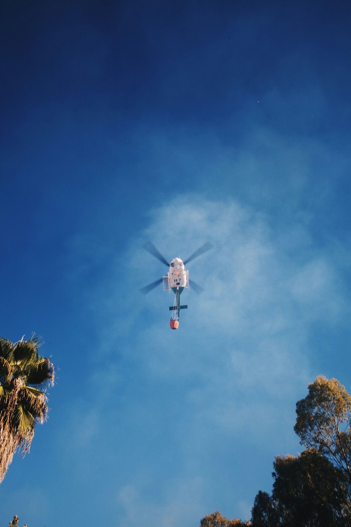 A helicopter seen from directly below against a clear blue sky, carrying a water bucket on a line beneath it.