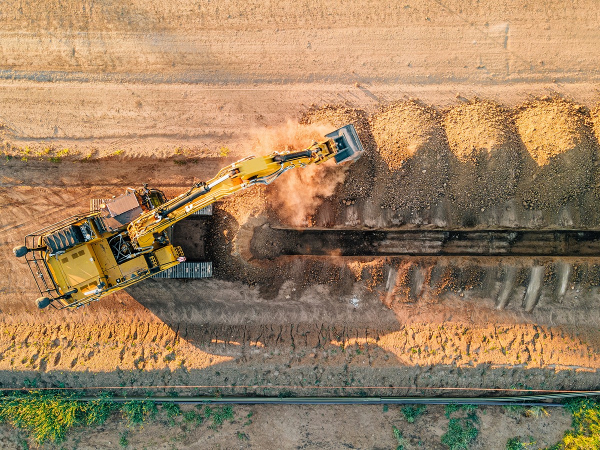 An overhead view of a yellow excavator cutting a trench through dry earth, spoil heaped along one side.