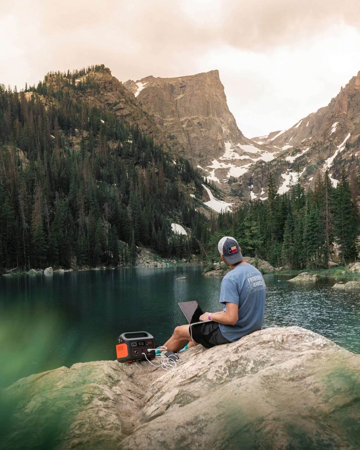 A person sits on a rock beside an alpine lake with a laptop, a small power station on the ground next to them, pine forest and a snow-streaked peak behind.