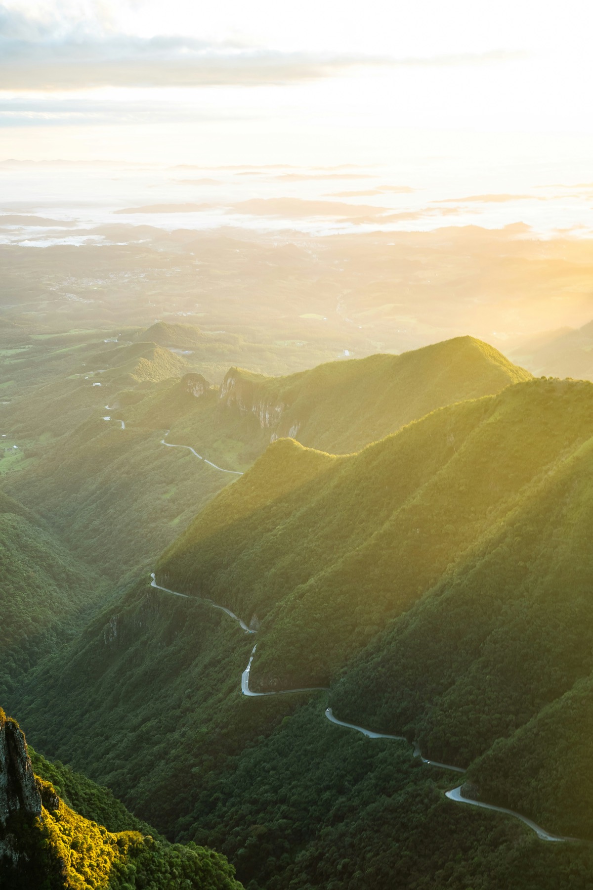 An aerial view of steep green forested ridges at sunrise, a narrow road winding along the valley floor in low golden haze.