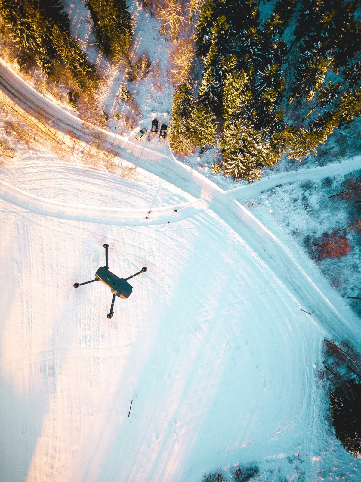 An aerial view looking straight down on a quadcopter drone flying over deep snow, with tracks, a road and dark conifers below.