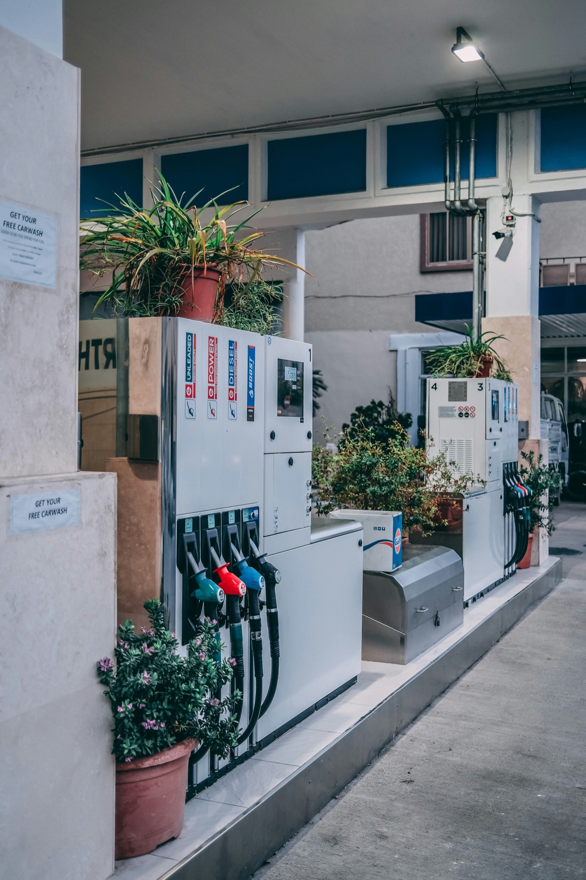 A small filling station forecourt with two fuel dispensers, nozzles in a row and potted plants along the wall behind.