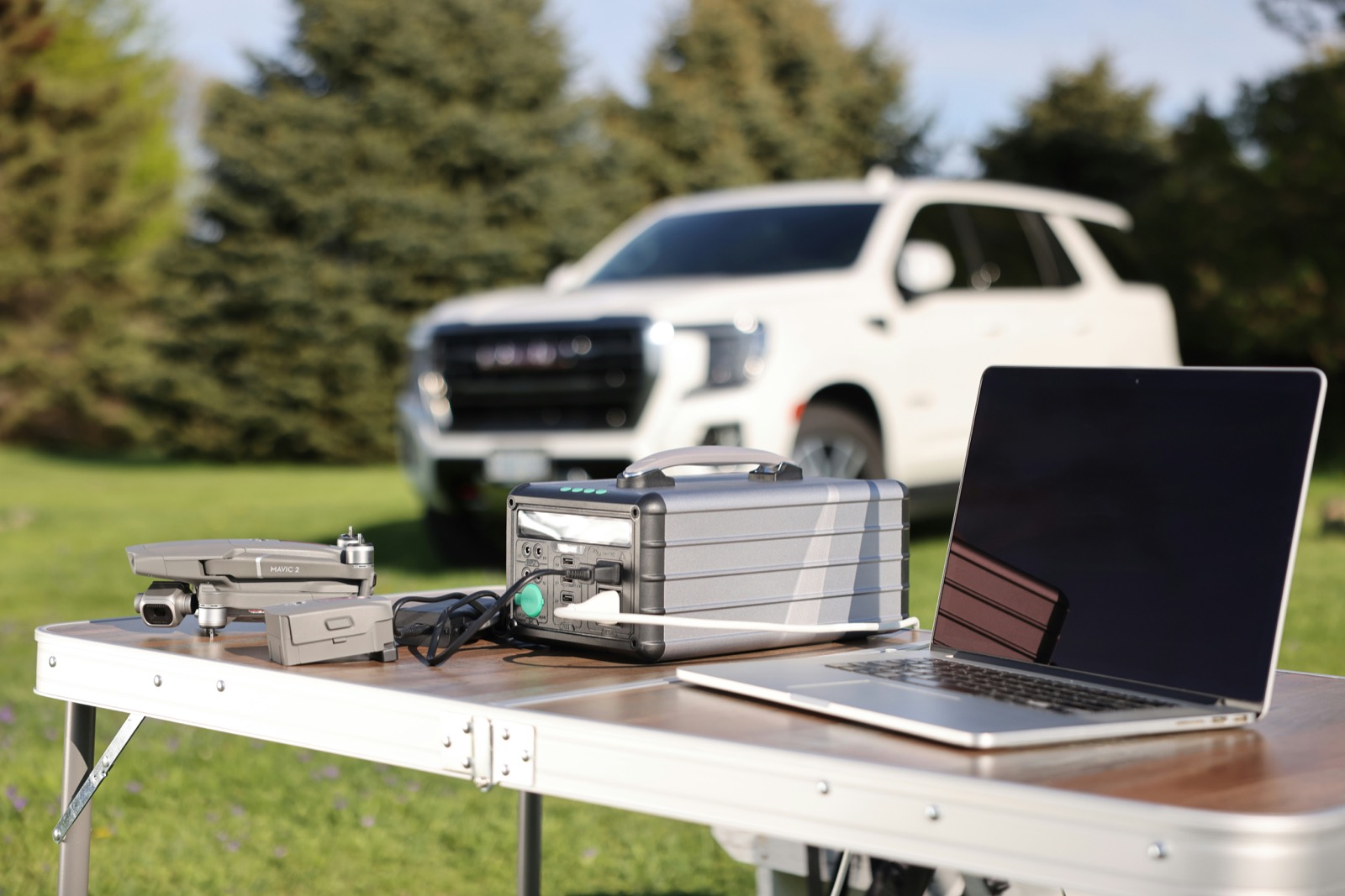 A portable power station on a folding camp table beside a laptop and a small drone, with a white pickup truck parked on grass behind.