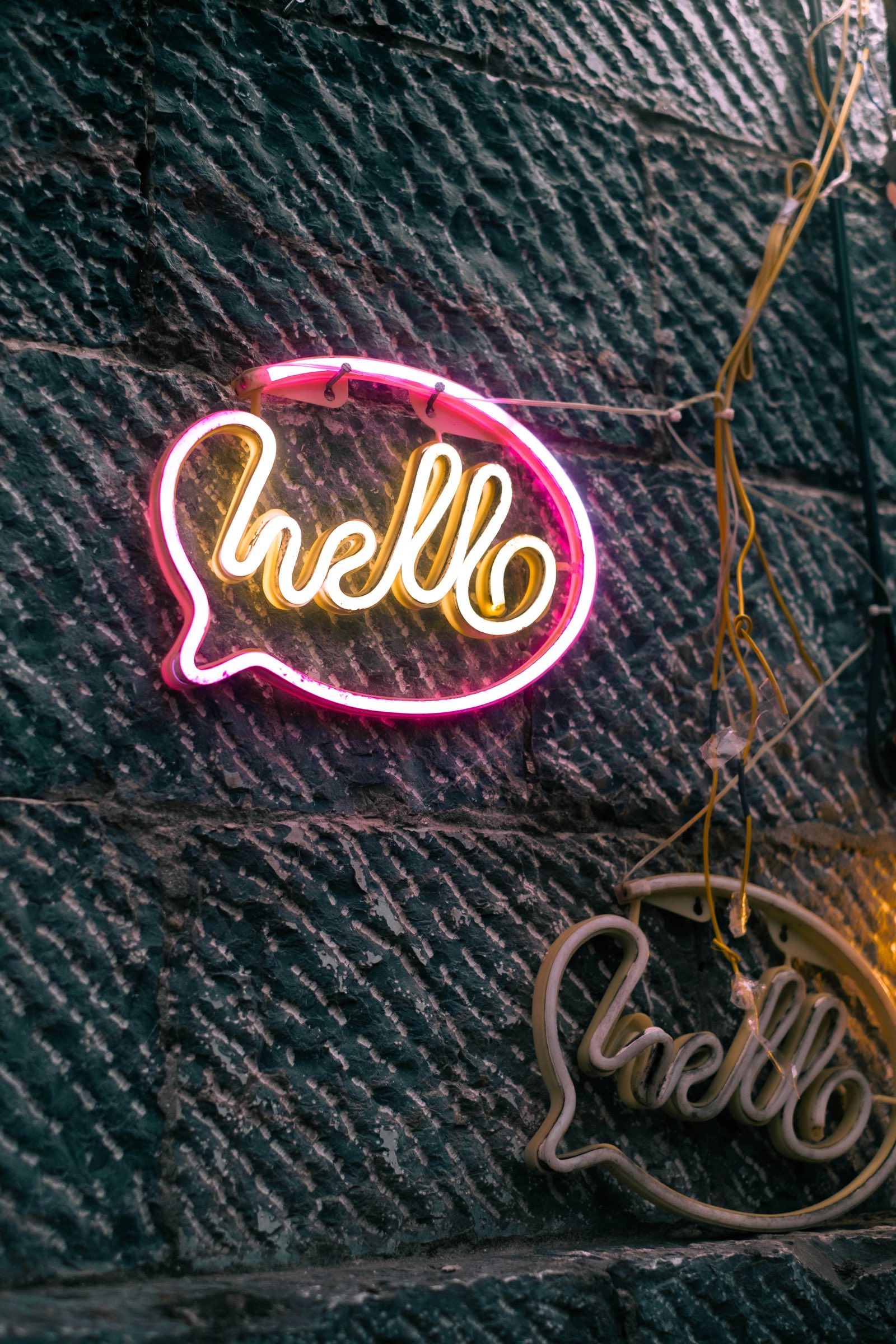 A neon sign shaped like a speech bubble glows pink and gold against a rough dark stone wall, spelling the word hello in looping script.