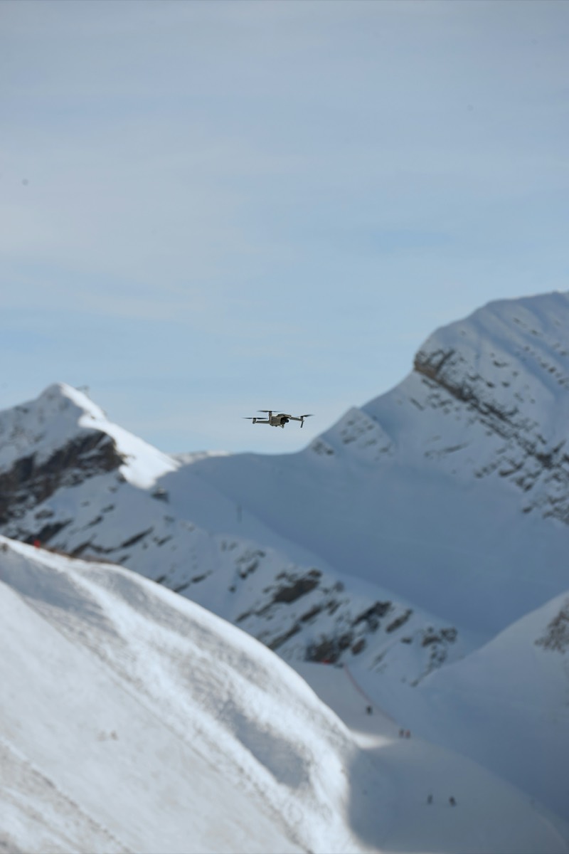A small quadcopter hovers above a snow-covered mountain ridge under a pale sky, with skiers on the slope far below.