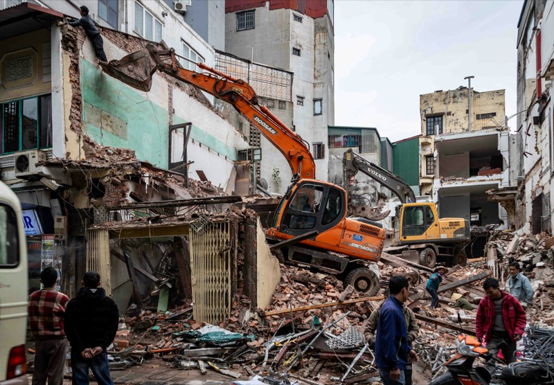 Two excavators pull down a partly demolished multi-story building while onlookers stand at the edge of the rubble in a narrow city street.