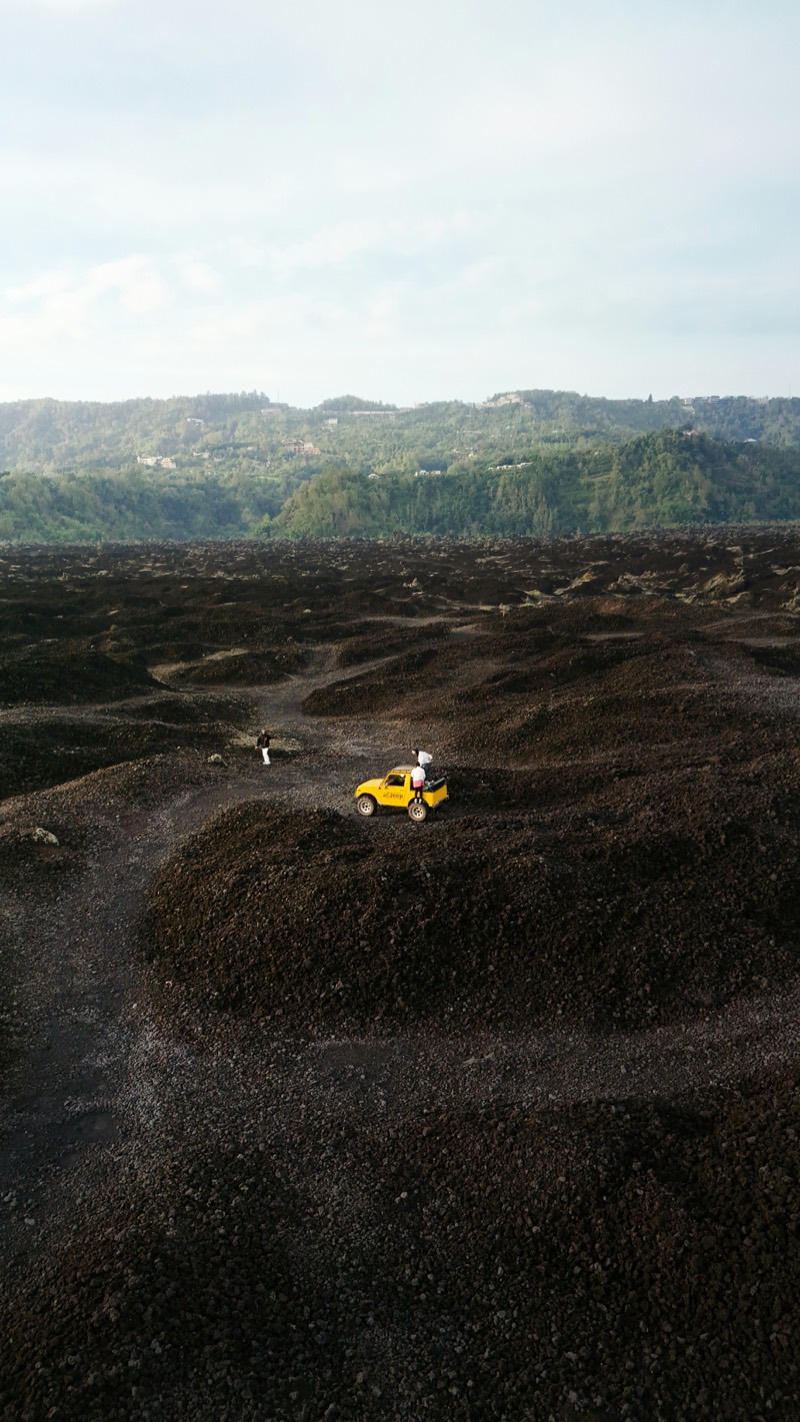 An aerial view of a yellow off-road buggy and two people crossing a wide field of black volcanic gravel, green forested hills behind.