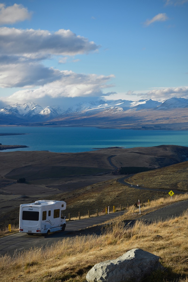 A white motorhome parked on a hillside road above a turquoise glacial lake, with a snow-capped mountain range on the far shore.