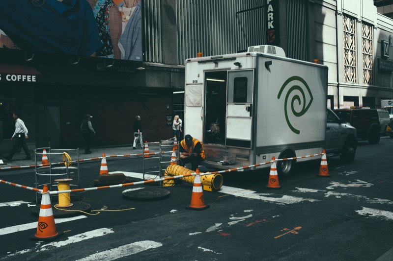 A utility crew works at an open manhole in a city street, with traffic cones, a ventilation duct and a box truck alongside.