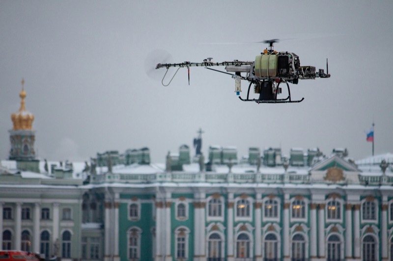 An unmanned single-rotor helicopter hovers above a river in front of a large baroque palace facade on a gray winter day.