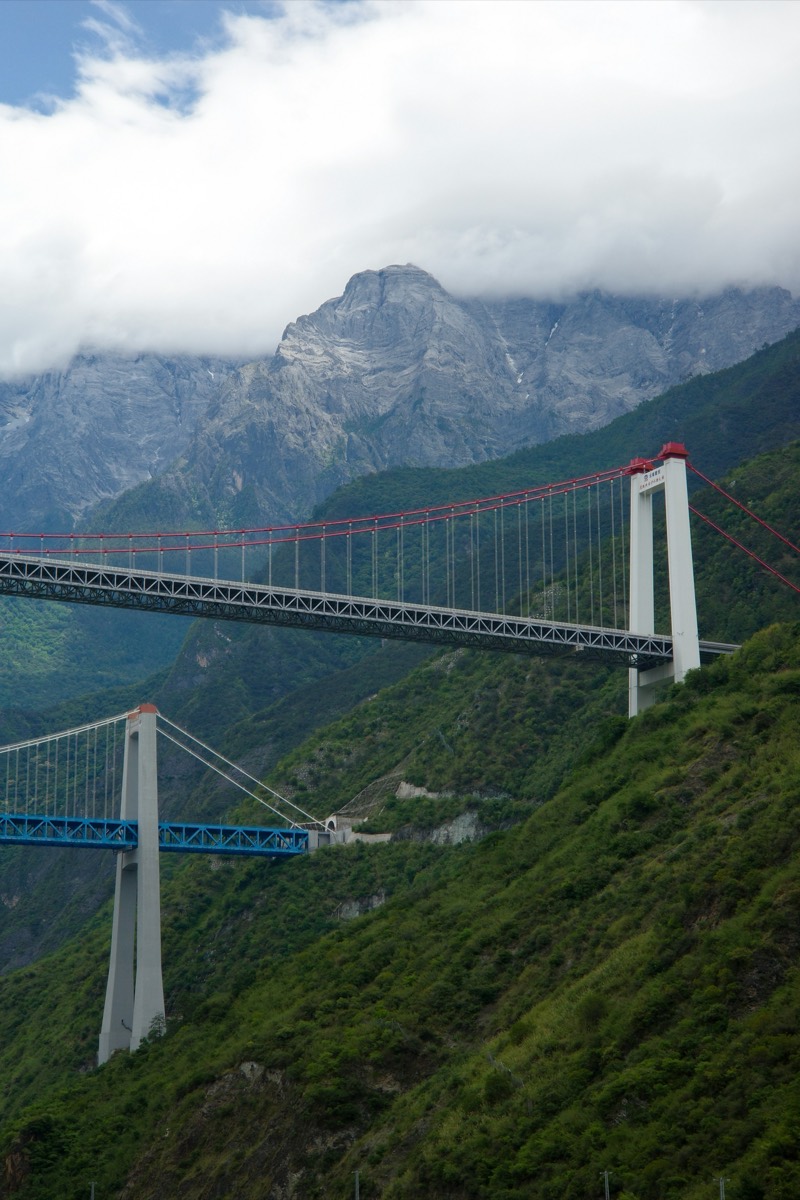 A suspension bridge with red and blue steel spans crosses a deep green gorge below a cloud-covered rock face.