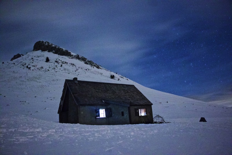 A small stone hut with lit windows stands alone in deep snow at night, below a rocky peak under a star-filled sky.